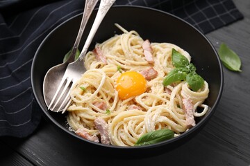 Bowl of delicious pasta Carbonara with egg yolk and cutlery on black wooden table, closeup