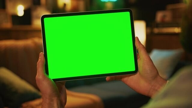 Shot of hands man holding and tapping on a tablet computer with a horizontal green screen. Mock up. Sofa on background