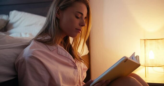 Blonde girl in a pink nightgown reads a book with a lamp on. Cozy book reading before going bed