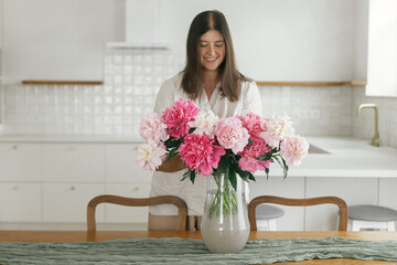 Woman holding beautiful peonies in vase on wooden table in new modern home. Young female decorating...