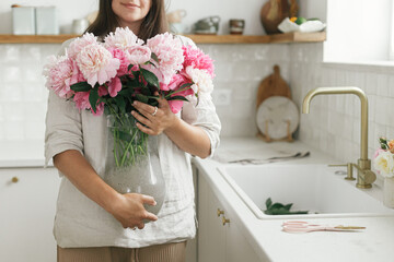 Woman holding beautiful peonies in vase at sink with brass faucet and granite countertop. Stylish female holding pink peony flowers in new modern kitchen, housewife decorating new home