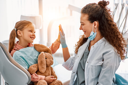 Cute little girl sitting in dentist chair, giving high five to female doctor and laughing. Dental care, trust and patient care. Children's dentistry.Sunlight.