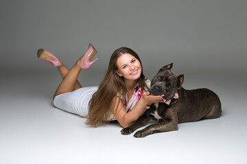 Obraz premium Beautiful elegant happy young woman in white dress hugging adult grey amstafford terrier dog with bow on neck. Studio shot over grey background. Copy space.