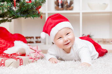 Cute baby boy in a Santa hat next to Christmas tree with presents
