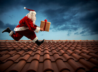Santa Claus runs with a big present on a house roof