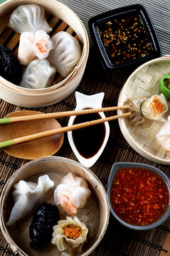 Arrangement of Various Dim Sum in Bamboo Steamed Bowls, Black and Red Chili Sauces, Soy Sauce and Chopsticks closeup on Straw Mat background. Top View