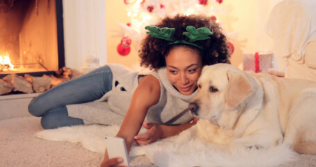 Cute young woman wearing a colorful green set of Christmas reindeer antlers taking a selfie with her dog as they relax in the living room in front of the fire.