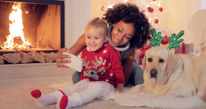 Happy family selfie portrait at Christmas with a happy smiling young woman photographing herself with a small girl and dog in reindeer antlers on the living room floor in front of a blazing fire.