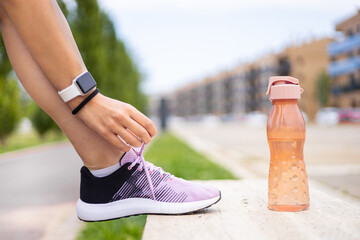 Sporty woman tying shoelaces on a bench at fitness training outdoors. Young female athlete running workout. Hydration, wellness, exercise, health and active lifestyle.