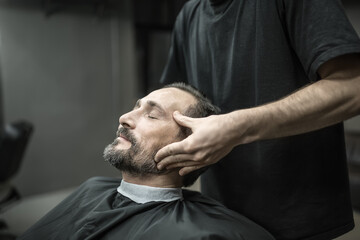 Handsome man with a beard and closed eyes in a black cutting hair cape in the barbershop. Barber in a black T-shirt is doing him a face massage. Closeup. Horizontal.