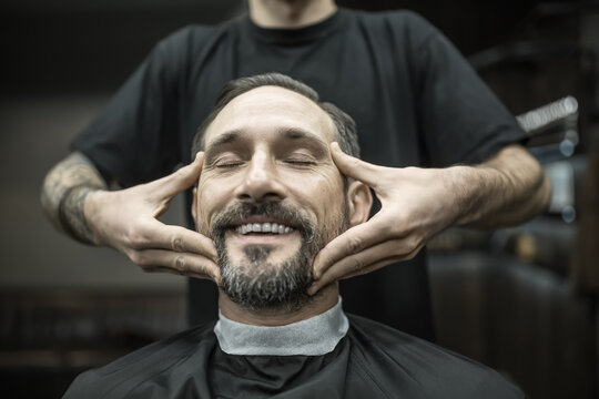 Smiling man with a beard and closed eyes in a black cutting hair cape in the barbershop. Barber with a tattoo in a black T-shirt is doing him a face massage. Closeup. Horizontal.