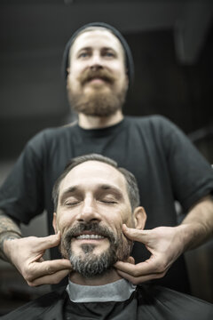 Pleased man with a beard and closed eyes in a black cutting hair cape in the barbershop. Bearded barber in a black T-shirt and a cap is doing him a face massage. Closeup. Vertical.