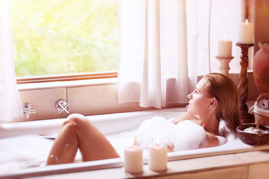 Beautiful woman lying down in the bath with foam and looking at the window, relaxation at home, hygiene and health care, happy and peaceful day spa