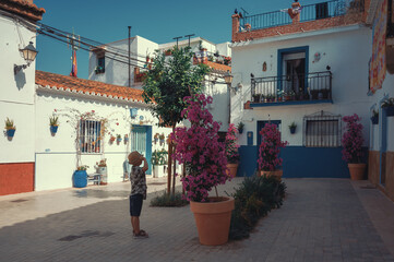 A boy tourist in a hat stands in front of bright flowers on a narrow street of the old town in Spain