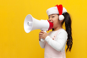 little asian girl in santa hat announces and shouts into megaphone on yellow background, korean...