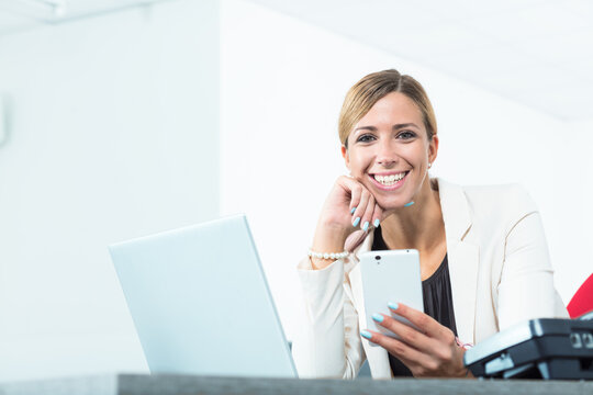 smiling woman holding a mobile phone as she works at her pc at the same time