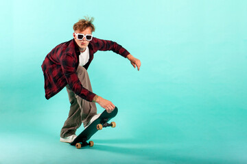 young cheerful guy rides skateboard on blue isolated background, hipster skater in sunglasses