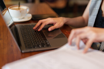 Close-up shot of Asian businesswoman hand morning of cafe sitting at the desk using a laptop computer and examined the documents lying on the table. Woman validating cafe account