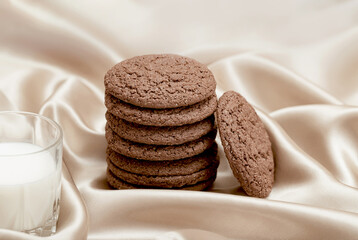Brown american cookie closeup with a glass of milk on beige silk fabric background. Minimal style. Bakery and pastry concept. Idea for breakfast.