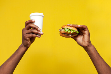african american man's hands hold delicious fresh burger and paper cup coffee on yellow isolated background - Powered by Adobe