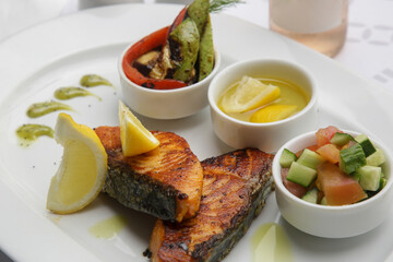 Fried salmon steak served with grilled vegetables, olive oil, lemon slices and pesto sauce on the restaurant table