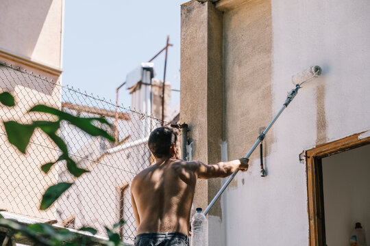 Unrecognizable shirtless young man painting wall with roller brush in daylight