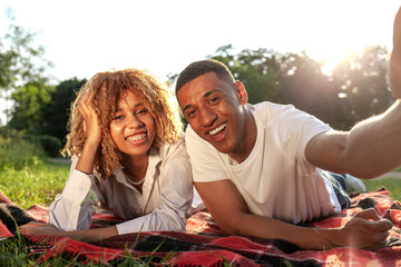 african american young couple lies in the park in the summer on the grass and takes selfie - obrazy, fototapety, plakaty