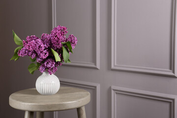 Beautiful lilac flowers in vase on wooden table near light wall, space for text