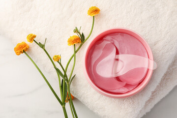 Jar of under eye patches with spoon, flowers and towel on white marble table, flat lay. Care...
