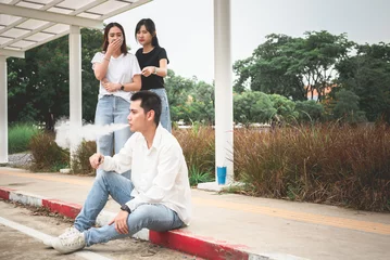 Foto op Canvas Asian man sitting on the sidewalk He is smoking e-cigarettes and spewed out a large amount of cigarette smoke, causing two women to pass by stink of cigarettes and feel disgusted. © Anatta_Tan