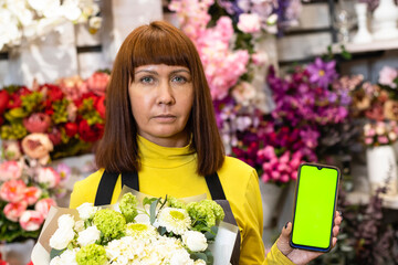 Floristry. Flowers. A florist girl with a bouquet of flowers, and with a phone with a green screen, chromakey. Shop, against the background of a showcase with garden decor.