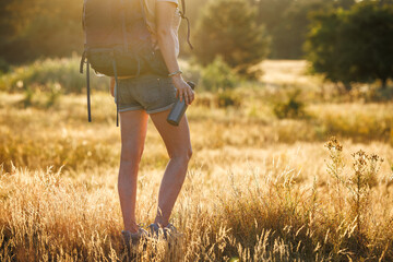 Woman with backpack and thermos is resting in nature and enjoying summer sunset. Relax while hiking on a long trekking trail