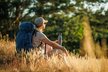 Naklejka na ściany i meble Woman with backpack is resting in forest and enjoying summer sunset. Relax while hiking on a long trek