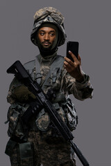 Dark skinned soldier in NATO uniform and helmet holds up his smartphone for a selfie or video chat on a plain grey background, showing the versatility and technology skills of military personnel