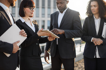 Crop of four multiracial colleagues in suits signing contract during business meeting on fresh air. African american corporate executive using e-signature on digital tablet with finger.