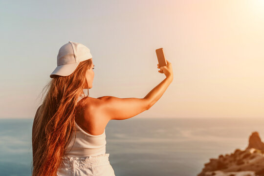 Selfie woman sea. The picture depicts a woman in a cap and tank top, taking a selfie shot with her mobile phone, showcasing her happy and carefree vacation mood against the beautiful sea background
