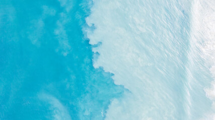 Top view of light blue water with white sand. Waves raise sand and a mix with turquoise water is obtained. Shadows from clouds on a clear ocean. Clear sea. Gradient of blue. The background photo