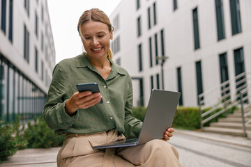 Smiling woman manager is using phone and work on laptop outside of office. Distance work concept