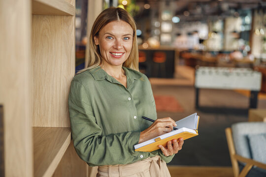 Pretty woman entrepreneur making notes in note pad while standing on cozy coworking background