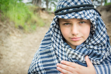 In the summer, a beautiful boy in a arafatka, an Arab scarf, stands on the road in the forest.