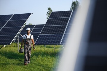 Portrait of young handsome African American craftsman in protective helmet. Man in uniform and with tools standing among solar panels
