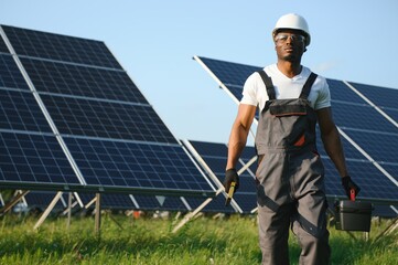 Portrait of young handsome African American craftsman in protective helmet. Man in uniform and with tools standing among solar panels