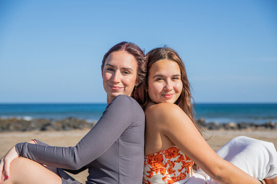 Two smiling sisters sitting back to back smiling and happy at the seaside