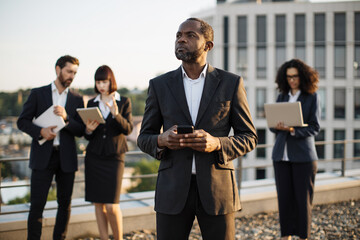 Good-looking corporate investor standing with smartphone in hands on terrace and controlling working process. Multicultural business partners dressed in official clothes using wireless devices.