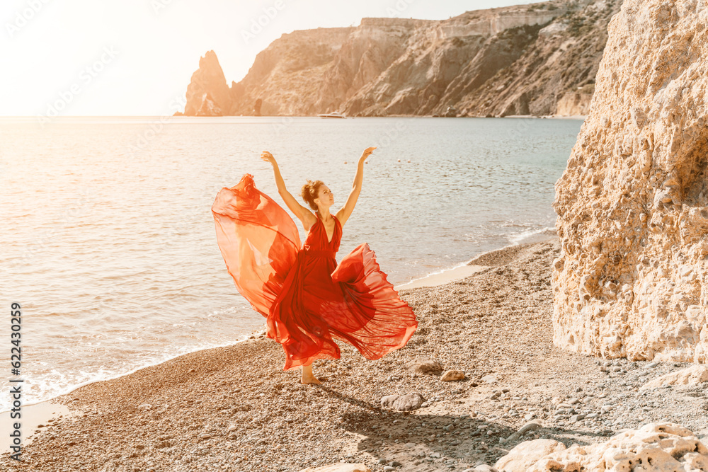 Wall mural Woman red dress sea. Female dancer in a long red dress posing on a beach with rocks on sunny day. Girl on the nature on blue sky background.