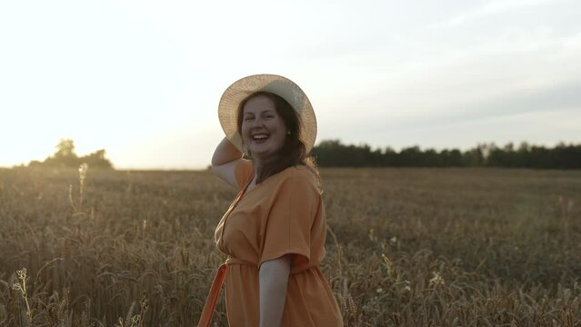 Happy middle aged woman in orange dress, hat twirls in wheat field, slow motion. Free plus size lady enjoys clean air, ecological area, smiling at camera. Concept of accepting self, carelessness.