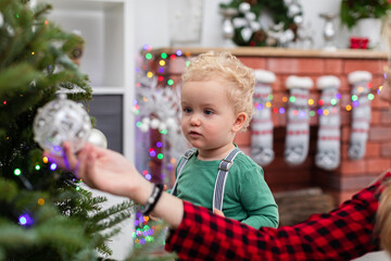 A little boy stands ip by the Christmas tree and watches the ornaments hanging on the branches.