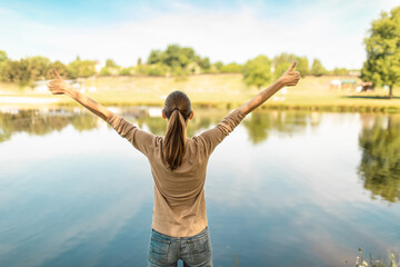Young adult woman raises arms, enjoying nature's beauty by tranquil lake with tree reflection.