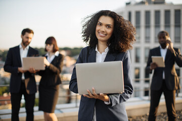 Portrait of pretty african american lady in business outfit posing on camera with laptop and smiling. Group of diverse entrepreneurs gathering on rooftop with gadgets to discuss new project plan.