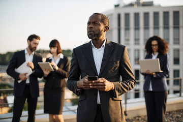 Good-looking corporate investor standing with smartphone in hands on terrace and controlling...